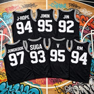 Könnte beinhalten: Sieben schwarze Basketballtrikots mit weißen Verzierungen und Schriftzügen werden gezeigt. Jedes Trikot hat einen anderen Namen und eine andere Nummer: J-HOPE 94, JIMIN 95, JIN 92, JUNGKOOK 97, SUGA 93, V 95 und RM 94. Die Trikots sind auf einem Basketballfeld angeordnet.