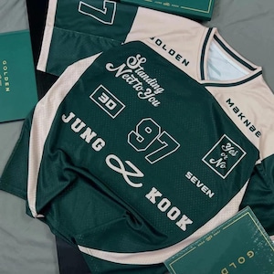 Puede incluir: Una camiseta verde oscuro y beige con letras y números blancos. La camiseta presenta el texto "Standing Next to You", "JUNG & KOOK", "97" y "Yes or No". La camiseta tiene un diseño de cuello en V.