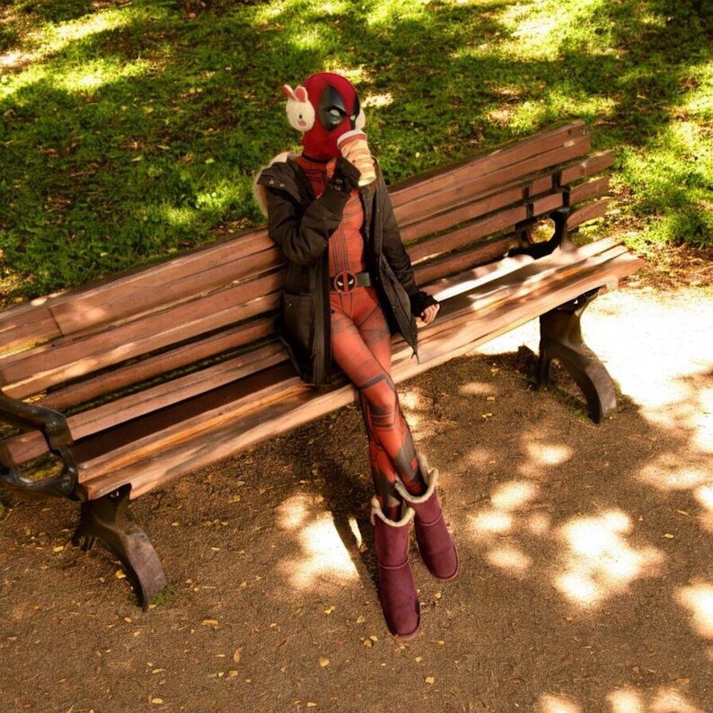 May include: A person wearing a Deadpool costume, including a red mask, sitting on a wooden park bench. They are wearing a black jacket, red and black patterned leggings, and burgundy boots. The person is holding a coffee cup.