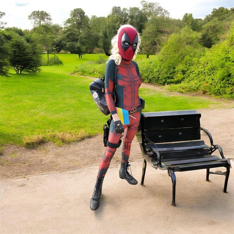 May include: A person in a Deadpool costume stands near a park bench. The costume is red and black with a mask. The person is wearing black boots and a backpack. A small flag with blue and yellow colors is visible.