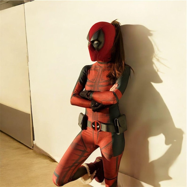 May include: A red and black Deadpool costume with a matching mask. The full-body suit features a belt with a Deadpool logo buckle. The person is leaning against a white wall.
