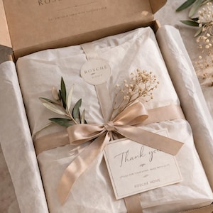Pode incluir: Uma caixa de presente da Rosche Home, cheia de linho. O conteúdo é embrulhado em papel de seda e amarrado com uma fita e laço bege. Inclui um raminho de vegetação e um cartão que diz "Thank you".