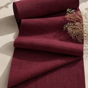 Pode incluir: Um caminho de mesa em tom bordô profundo feito de tecido entrelaçado com uma textura visível semelhante ao linho. O caminho está parcialmente enrolado no topo e drapeado para baixo em dobras, com uma bainha simples nas laterais e base.