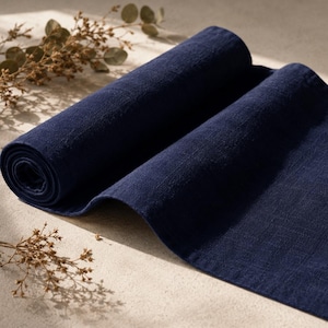 Op de afbeelding: Een donkerblauwe stoffen loper met een zichtbare geweven textuur, gedeeltelijk opgerold. Het textiel heeft een consistente diepblauwe tint over het gehele oppervlak en toont een eenvoudige, rechthoekige vorm met strakke, afgewerkte randen.
