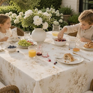 Op de afbeelding: Een tafel gedekt voor een maaltijd in de buitenlucht, met een bloemig tafelkleed, wit servies en een vaas met witte bloemen. De tafel is gedekt met fruit, sap en croissants. Twee kinderen zitten aan tafel.