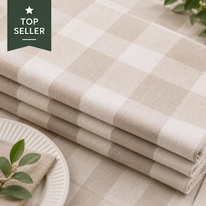 May include: A folded beige and white checkered tablecloth with a textured surface. A tag attached to the fabric displays the text: WATERPROOF Repels spills & liquids, EASY TO CLEAN Wipe clean in seconds, DURABLE Built to last. A top seller label is visible.