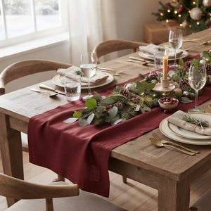 May include: A wooden dining table set for a festive meal with a deep red table runner, white plates with gold rims, beige linen napkins, gold-toned cutlery, clear glassware, and a centrepiece of greenery, pinecones, and lit taper candles.