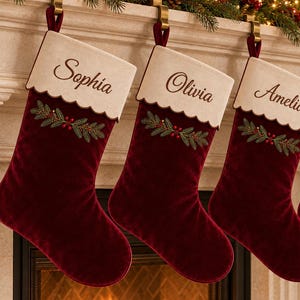 Op de afbeelding: Vier dieprode kerstsokken van fluweel hangen aan een schoorsteenmantel. Elke sok heeft een crèmekleurige boord met een geschulpte rand, geborduurd met de namen Sophia, Olivia, Amelia en James, en een takje met hulstbessen.