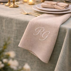 May include: A beige linen napkin with embroidered white initials, laid on a table set for a formal dinner. The table has a light green tablecloth and is decorated with gold cutlery, candles, and a floral centrepiece.