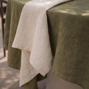 May include: A linen tablecloth in olive green covers a round table, with a folded natural linen napkin draped across it. The table setting is simple and elegant, suitable for outdoor dining.