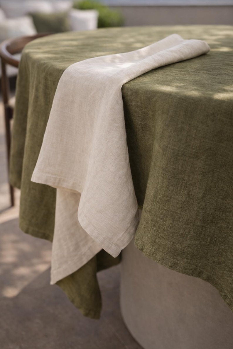 May include: A linen tablecloth in olive green covers a round table, with a folded natural linen napkin draped across it. The table setting is simple and elegant, suitable for outdoor dining.