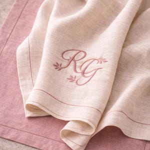 May include: A folded beige linen napkin with embroidered initials "RG" in a script font, accented with floral details. The napkin rests on a pink placemat, with a stitched border. The overall aesthetic is soft and elegant.