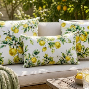 Pode incluir: Quatro almofadas decorativas com um padrão de limão e folhas sobre um fundo branco. As almofadas estão dispostas num sofá bege. Uma manta verde está sobre o sofá. Uma mesa de madeira tem uma tigela com limões e um copo de limonada.