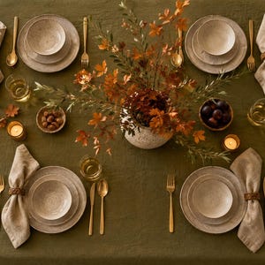 Könnte beinhalten: Ein gedeckter Tisch mit olivgrüner Tischdecke, vier Gedecken mit gesprenkelten beigen Tellern und Schalen, goldenem Besteck, Leinentüchern in geflochtenen Ringen, klaren Gläsern, kleinen Kerzen und einer zentralen Vase mit herbstlichem Laub.