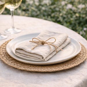 May include: A folded beige linen napkin tied with twine sits on a white plate, atop a woven placemat. The setting is on a white tablecloth, with a blurred background of greenery and wine glasses.