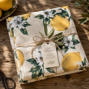 Könnte beinhalten: Eine gefaltete Leinentischdecke mit einem Muster aus gelben Zitronen, weißen Blüten und grünen Blättern. Der Stoff ist mit einer Schnur zusammengebunden und trägt ein Etikett mit der Aufschrift Linen Tablecloth, 100% Linen, MADE IN EUROPE, und einem Blatt.