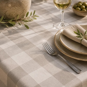 Peut inclure: Une nappe à carreaux beige et blanc avec une texture tissée. Le couvert comprend une pile de deux assiettes en céramique mouchetée, une serviette en lin beige pliée avec un petit brin vert, une fourchette en argent, un verre à vin et un bol d'olives.