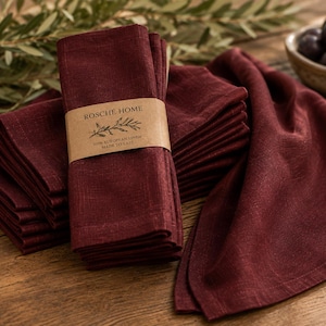 May include: A stack of deep burgundy linen napkins wrapped in a tan paper band labelled ROSCHÉ HOME 100% EUROPEAN LINEN MADE TO LAST. The napkins have a visible woven texture and are folded neatly, with one napkin draped to the side.