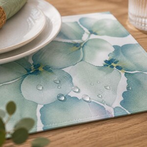 Pode incluir: Um jogo americano quadrado com um design floral em aquarela em tons de verde e azul. Gotas de água são visíveis no jogo americano. Pratos brancos e um guardanapo verde com um porta-guardanapo tecido estão no fundo.