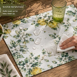 Pode incluir: Um jogo americano branco resistente à água com um design de limão e floral. O jogo americano tem um padrão de limões amarelos e flores brancas com folhas verdes. O texto "WATER RESISTANT" é visível. Uma mão limpa gotas de água do jogo americano.