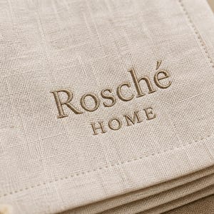 Può includere: Primo piano di una pila di tovaglioli di lino beige con il testo ricamato "Rosché HOME" in filo marrone abbinato. I tovaglioli hanno un aspetto strutturato e un bordo cucito.