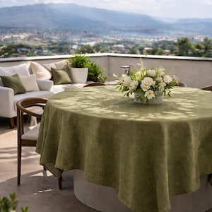 Può includere: Un tavolo da pranzo rotondo all'aperto con una tovaglia di lino verde. Una composizione floreale bianca è al centro. Il tavolo è circondato da sedie e un divano bianco con cuscini verdi. Un grande ombrellone fornisce ombra.