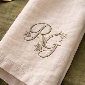 May include: Beige linen napkin with embroidered initials "RG" in olive green. The initials are embellished with delicate leaf designs, creating an elegant and personalized touch. The napkin is folded, showcasing its texture and quality, perfect for a formal dining setting.