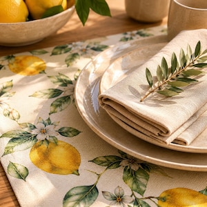May include: A table setting featuring a beige linen napkin and plate, adorned with a sprig of green leaves. The table runner displays a lemon and floral pattern. A bowl of lemons is visible.