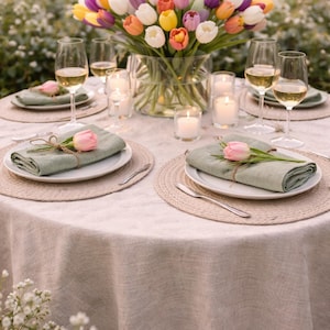 May include: A table setting with a bouquet of colorful tulips in a glass vase. The table is set with white plates, green napkins, and wine glasses. Candles and place settings are arranged on a beige tablecloth, creating an elegant outdoor dining scene.