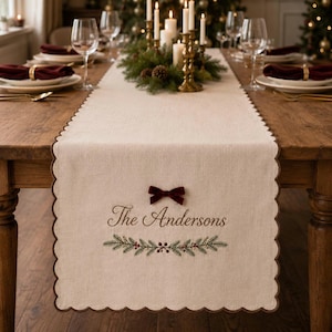 May include: A beige linen table runner with scalloped edges, featuring the embroidered text The Andersons, a small dark red velvet bow, and a green pine branch motif with red berries centred on the fabric surface.