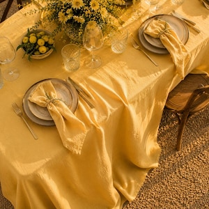 Könnte beinhalten: Ein schön gedeckter Tisch mit gelber Tischdecke, Tellern, Servietten und Besteck. Der Tisch ist mit einem Blumengesteck und einer Schale mit gelben Früchten geschmückt. Weingläser und Wassergläser sind ebenfalls vorhanden, was eine einladende Essszene schafft.