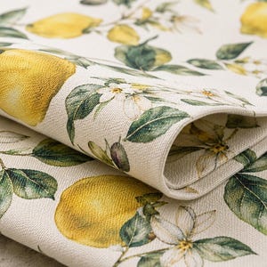 May include: Close-up of a folded linen fabric featuring a vibrant lemon and floral pattern. The design includes large yellow lemons, green leaves, and delicate white blossoms against a cream-colored background. The fabric has a textured appearance.