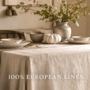 以下が含まれることがあります： 100% EUROPEAN LINENという文字がプリントされたベージュのリネン製テーブルクロス。テーブルには、白い陶器の皿2枚、お揃いのボウル2つ、白い装飾用カボチャ、緑の葉が入った灰色の花瓶が置かれています。