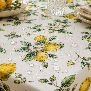 Può includere: Una tovaglia bianca con un motivo a limoni e fiori. Il design presenta limoni gialli, foglie verdi e fiori bianchi. Goccioline d'acqua sono sparse sulla superficie, suggerendo un materiale impermeabile o idrorepellente.