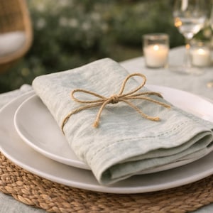 May include: A table setting with a light blue linen napkin tied with twine, placed on a white plate. The plate rests on a woven placemat. Wine glasses, candles, and gold-colored cutlery are also visible.