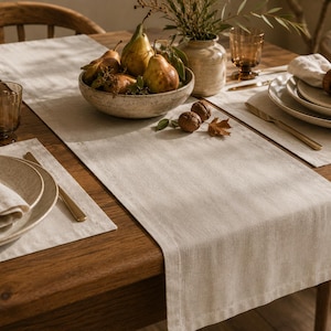 Op de afbeelding: Een lichtbeige linnen tafelloper op een houten tafel, vergezeld van bijpassende placemats, witte stoffen servetten met houten ringen, keramische borden, amberkleurig glaswerk, goudkleurig bestek en een schaal met peren.