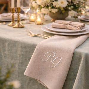 May include: A beige linen napkin with embroidered white initials "RG" rests on a sage green tablecloth. The table is set with gold cutlery, white plates, and a floral centerpiece. Lit candles and candle holders add to the ambiance.
