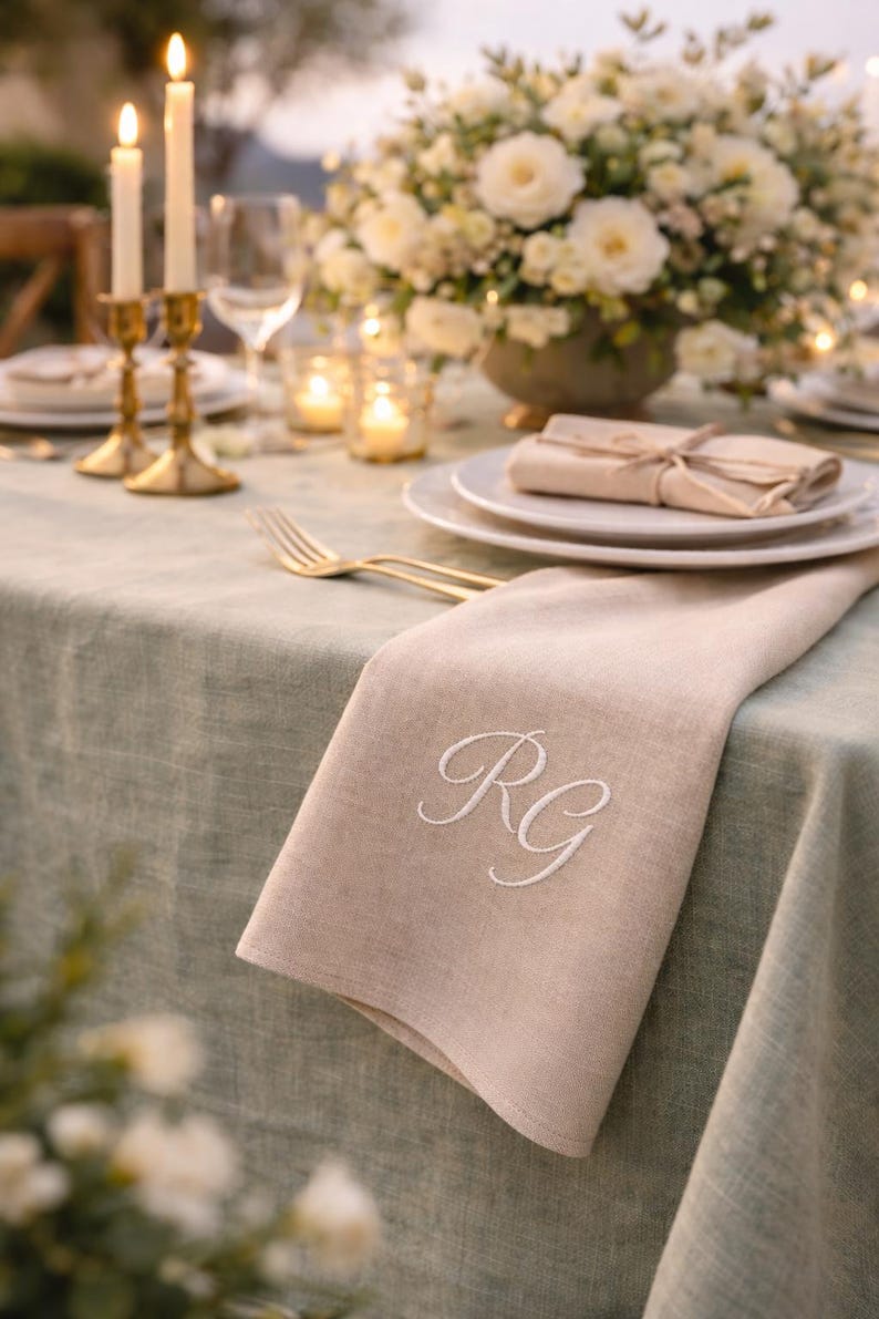 May include: A beige linen napkin with embroidered white initials "RG" rests on a sage green tablecloth. The table is set with gold cutlery, white plates, and a floral centerpiece. Lit candles and candle holders add to the ambiance.