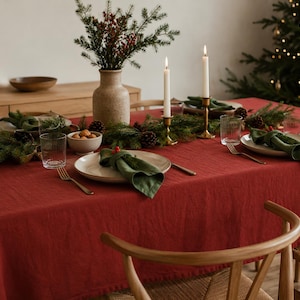 Puede incluir: Un mantel rojo liso que cubre una mesa de comedor con platos blancos, servilletas de tela verde, vasos transparentes, tenedores dorados y dos velas blancas en candelabros de latón, decorado con un centro de ramas de pino y piñas.