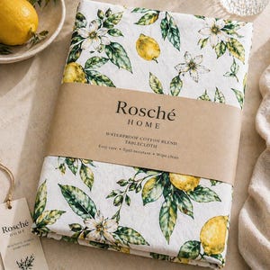 May include: A folded white tablecloth with a lemon and floral pattern. The tablecloth is wrapped with a brown paper band that reads "Rosché HOME WATERPROOF COTTON BLEND TABLECLOTH". A tag with the "Rosché HOME" logo is attached. A lemon and a bowl are in the background.