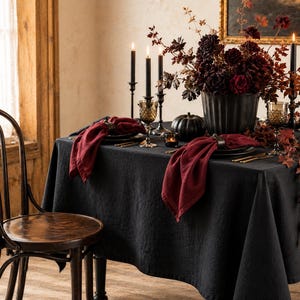 May include: A dining table set with a black tablecloth, dark red cloth napkins, and black plates. The centerpiece features a black vase with dark red flowers, black taper candles in holders, and a small black pumpkin, accompanied by a wooden chair.