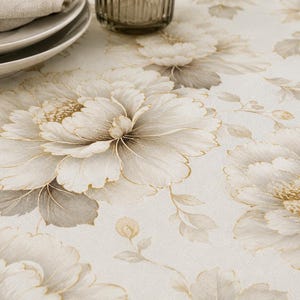 Puede incluir: Primer plano de un mantel floral con grandes flores de peonía color crema con detalles dorados. Las flores se destacan sobre un fondo beige claro, creando una estética suave y elegante. El mantel es adecuado para comer.