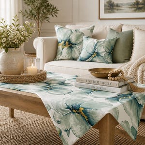 May include: A living room scene featuring a light-colored sofa with floral pillows and a matching table runner. A woven tray holds a vase of flowers, a candle, and a glass. A gold bowl and books are on the table.