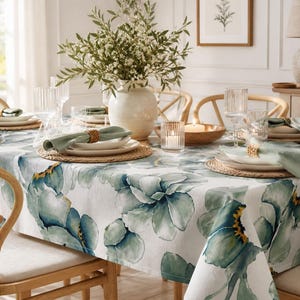 May include: A dining table set for a meal, featuring a white tablecloth with large blue and green floral patterns. The table is set with plates, napkins, glasses, and a vase of flowers. Wicker placemats and wooden chairs complete the scene.