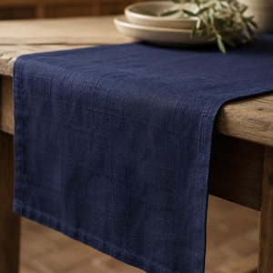 Pode incluir: Um caminho de mesa retangular azul-marinho feito de um tecido entrelaçado com uma textura visível. O caminho apresenta bordas retas e bainhas, e está colocado sobre a superfície de uma mesa de madeira, exibindo uma cor sólida e profunda.