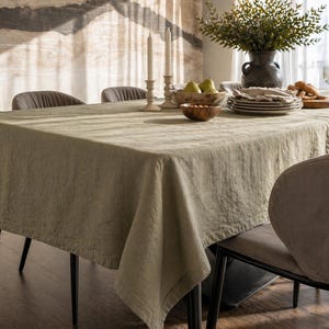 Könnte beinhalten: Eine salbeigrüne Leinentischdecke bedeckt einen Esstisch mit schwarzen Beinen. Der Tisch ist mit Tellern, Schalen, Kerzen und einer Vase mit Grünzeug gedeckt. Die Stühle sind neutralgrau.