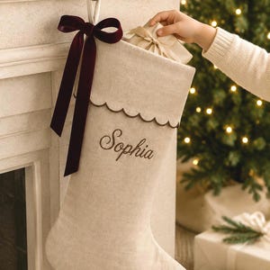 May include: A beige linen Christmas stocking featuring a scalloped top edge with dark brown trim and a deep burgundy velvet ribbon bow. The name Sophia is embroidered in dark brown script on the front of the stocking, which hangs from a hook.