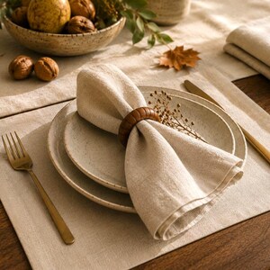 Peut inclure: Une serviette en lin beige pliée dans un rond de serviette en bois brun foncé, posée sur un ensemble d'assiettes en céramique mouchetée. Une fourchette et un couteau dorés sont disposés sur un set de table en lin beige assorti.