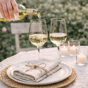 May include: A bottle of white wine being poured into a wine glass. Two wine glasses are filled with wine. A white plate with a folded linen napkin tied with twine sits on a woven placemat. Lit candles in glass holders are on the table.