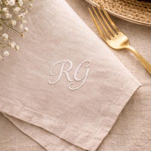 May include: Beige linen napkin with embroidered white initials "RG". A gold fork rests on the napkin, next to a woven plate. White flowers are in the upper left corner. The napkin is folded and has a textured appearance.